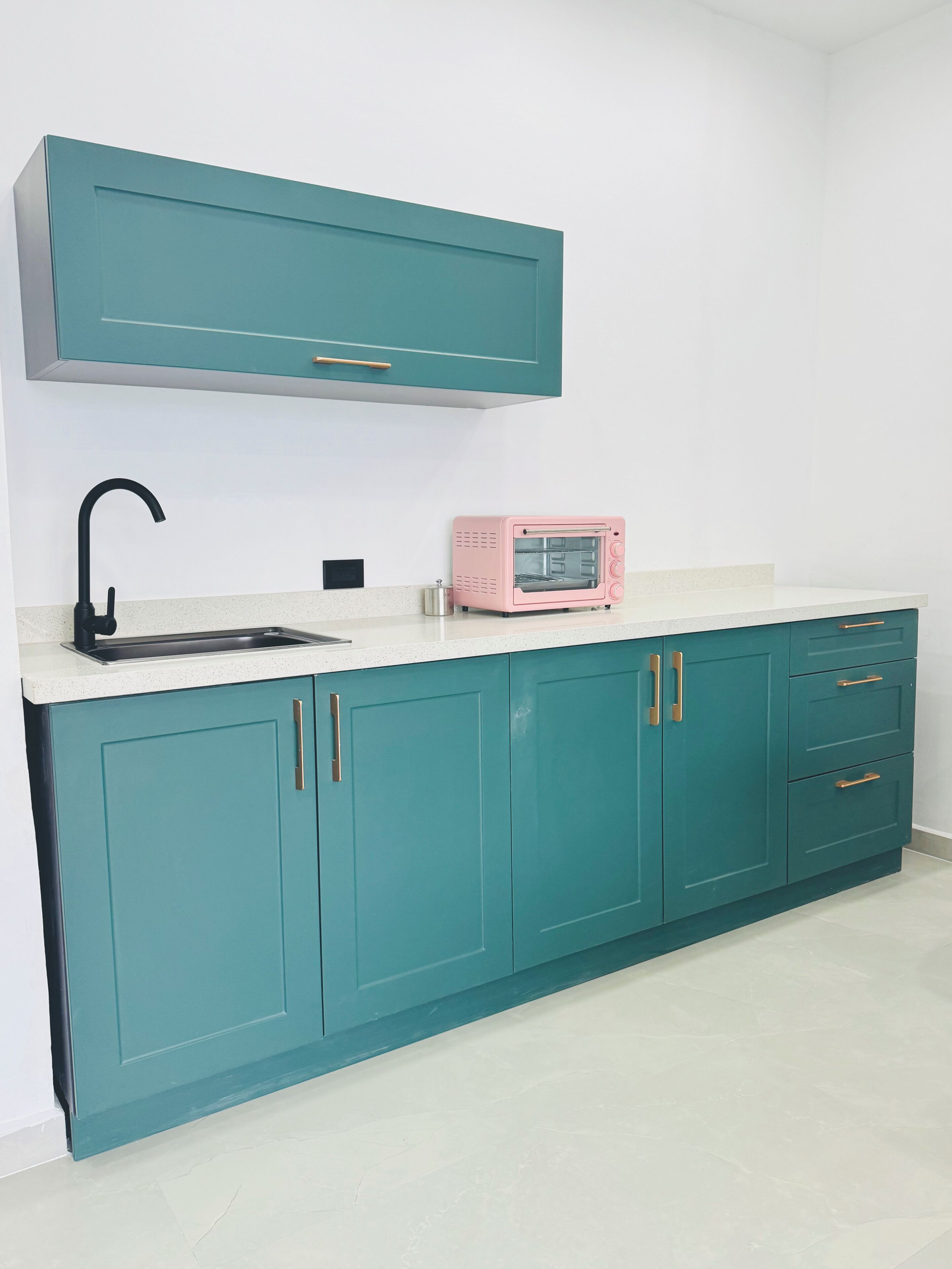 A kitchen with teal cabinets, a white countertop, a black faucet, and a pink toaster oven.
