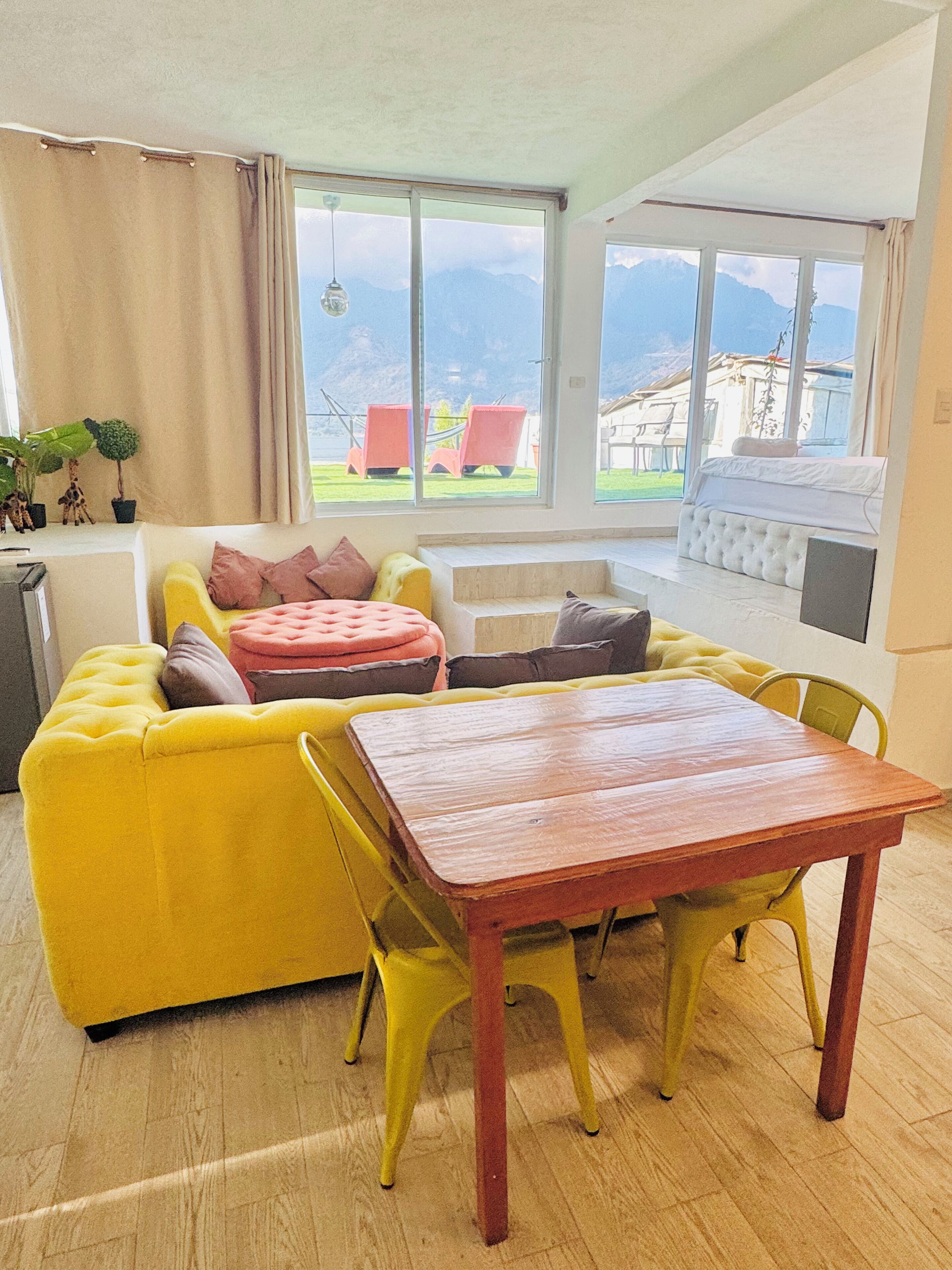 Una habitación luminosa con un sofá amarillo, una mesa de madera y vistas a montañas a través de amplios ventanales.