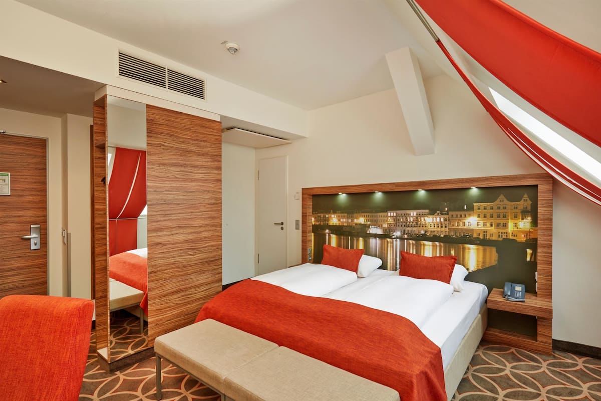 A hotel room with a bed, a bench, a nightstand, a mirror, and a cityscape mural on the wall.