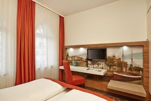 A hotel room with a bed, a desk with a chair, a television, and a cityscape painting on the wall. - H+ Hotel Lübeck (Lubeck)