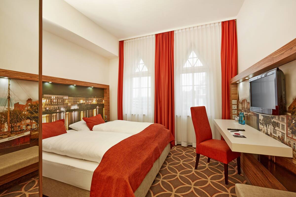 A hotel room with a bed, a desk, a chair, a television, and a large window with curtains.