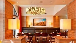 A restaurant with a central seating area, tables, and chairs, and a large painting on the wall. - H+ Hotel Lübeck (Lubeck)