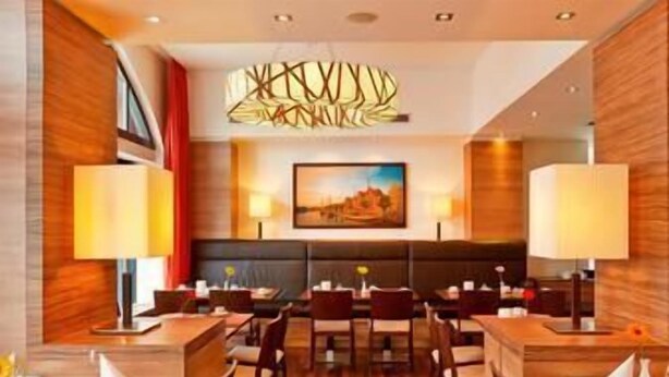 A restaurant with a central seating area, tables, and chairs, and a large painting on the wall. - H+ Hotel Lübeck (Lubeck)