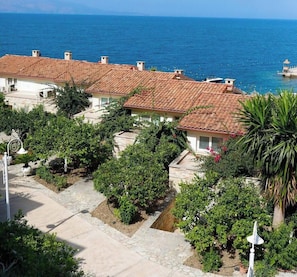 Zona residencial costera con casas de techo de tejas rojizas, un mar azul cristalino y un camino que lleva a la orilla.