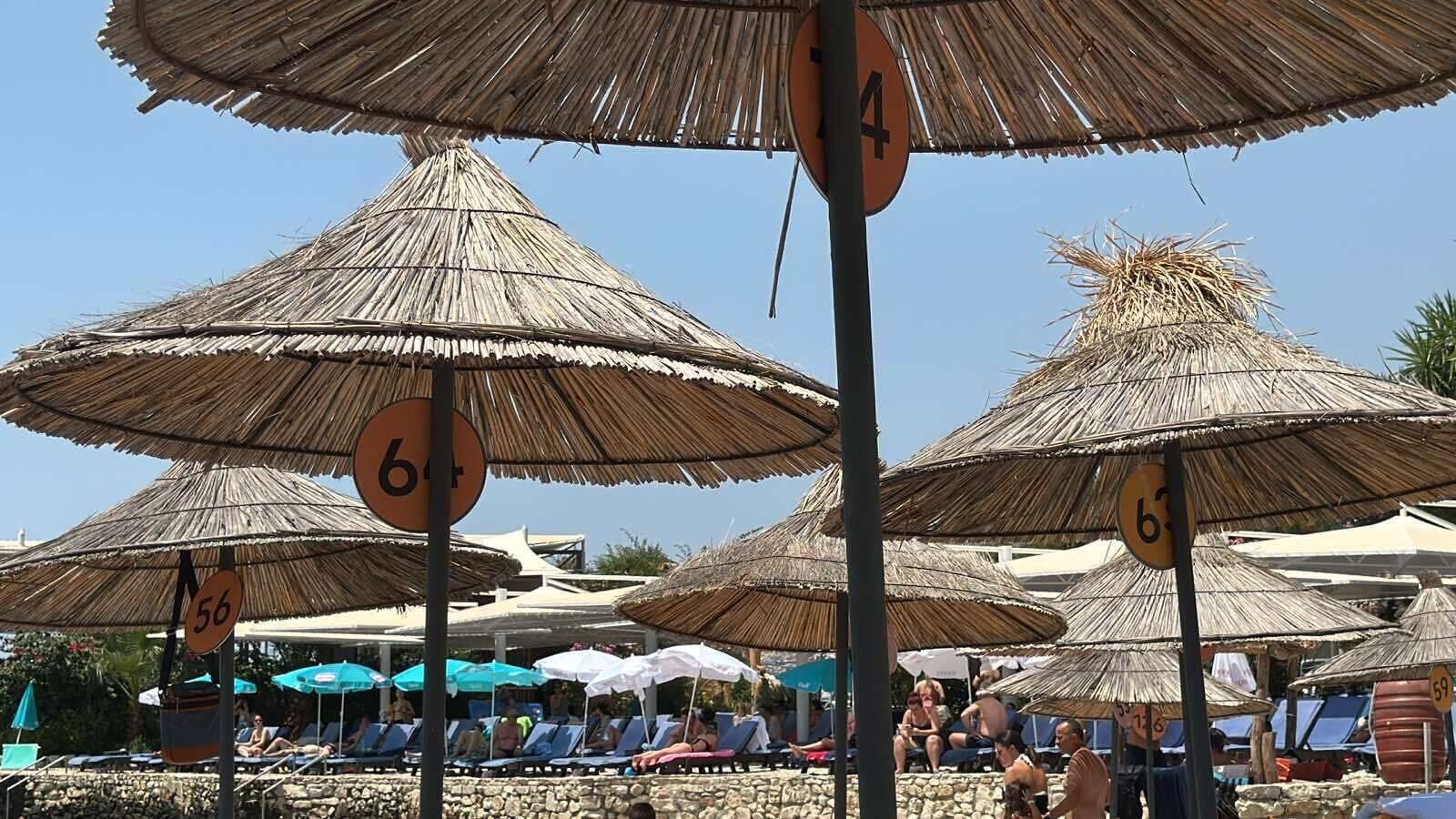 Beach umbrellas with numbers indicating their positions, a stone-paved area, and people relaxing under the umbrellas.