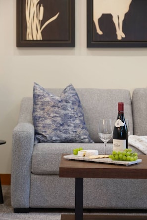 A living room with a grey sofa, a blue patterned pillow, a wooden coffee table with a bottle of wine, a glass, and a cheese board with grapes. - 2BR/2BA Steps to Olympic Rings! (Whistler)