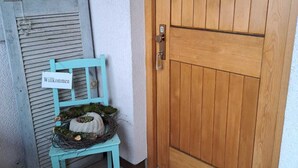 Une porte en bois avec une poignée en métal, une chaise bleu clair ornée d’une pancarte indiquant « Willkommen », et une petite couronne posée sur la chaise.