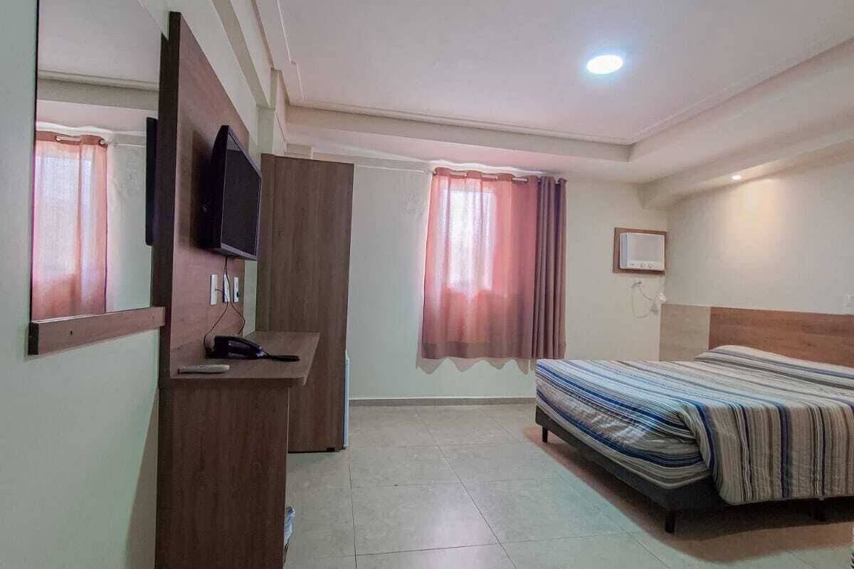 A bedroom with a bed, a desk, a TV, and a window with curtains.