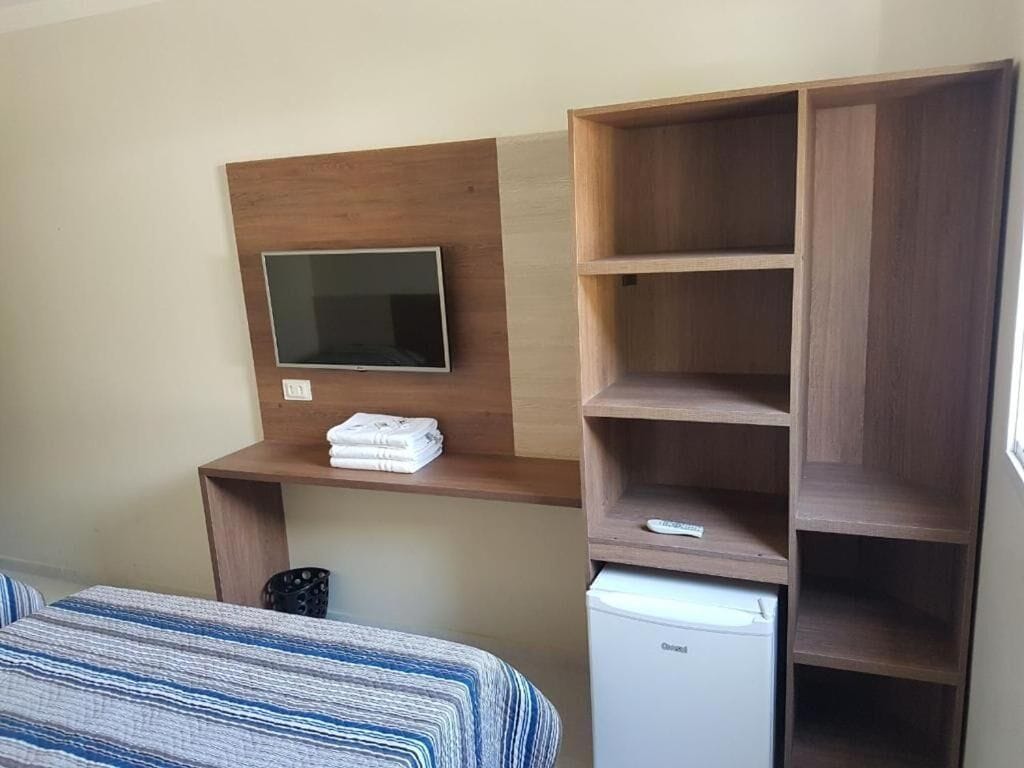 A hotel room with a bed, a TV mounted on a wooden shelf, a small refrigerator, and a wooden wardrobe.