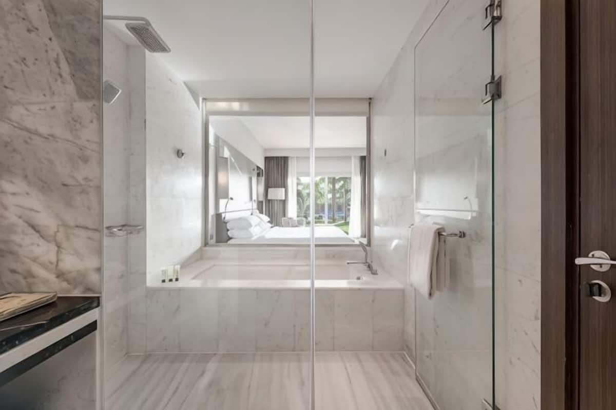 A modern bathroom with a glass shower enclosure, a marble floor, and a view into the adjacent bedroom.
