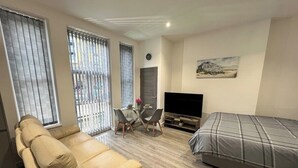 Een moderne woonkamer met een beige bank, een grijs bed, een televisie en een eethoek met een tafel en stoelen.