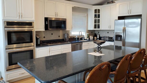 A modern kitchen with white cabinets, stainless steel appliances, a central island, and a dining area with wicker chairs. - Ship Bottom 5 BR Ocean block -Pool (Ship Bottom)