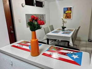 A dining area with a table set for two, a vase with red flowers, and a painting on the wall. - Condado Beachfront apartment with pool access (San Juan)