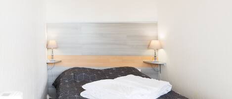 En lille, pænt seng med et mønstret sengetæppe og et hvidt dyne, to sengeborde med lamper og en lys farvet sengegavl.