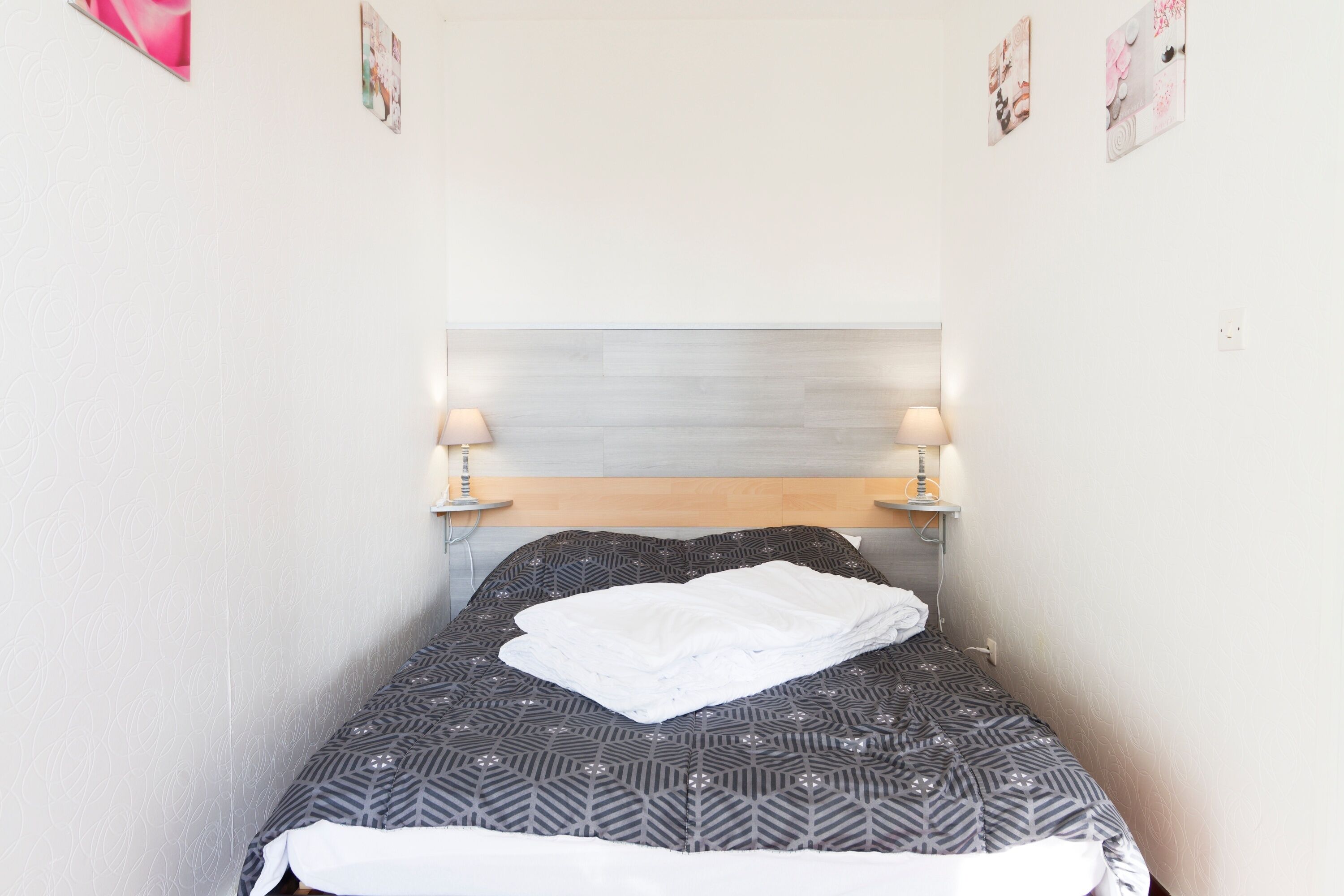 Uma cama pequena e bem arrumada, com colcha estampada e manta branca, duas luminárias de cabeceira e cabeceira de madeira.