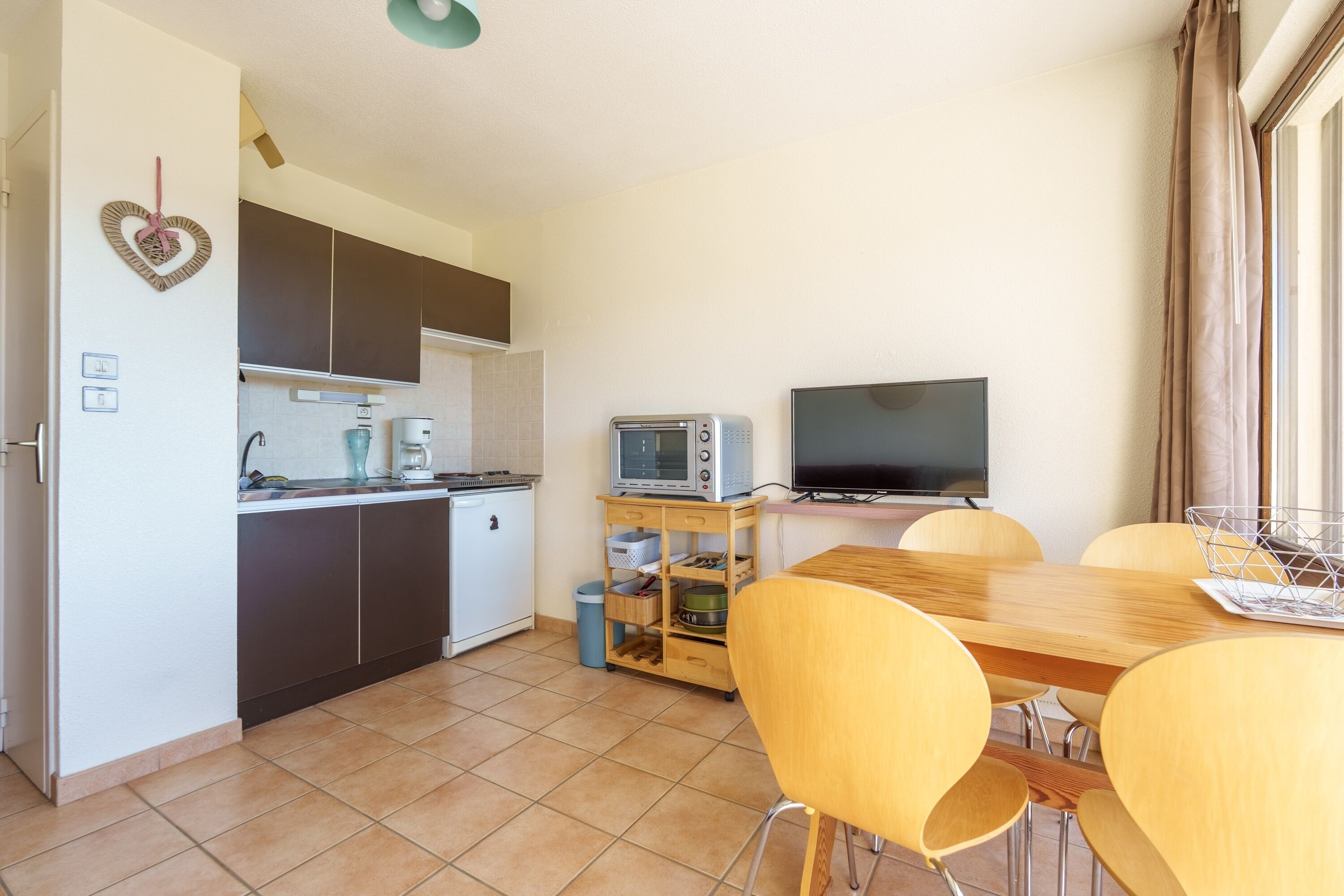 A compact kitchen with a microwave, toaster, and a flat-screen TV. There is a dining table with chairs and a small trash bin.