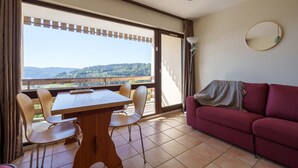 A room with a wooden table and chairs, a red sofa, a large window with a view of hills, and a round mirror on the wall. - Le Cocon du Chaud Costet - Avec balcon (Le Tholy)