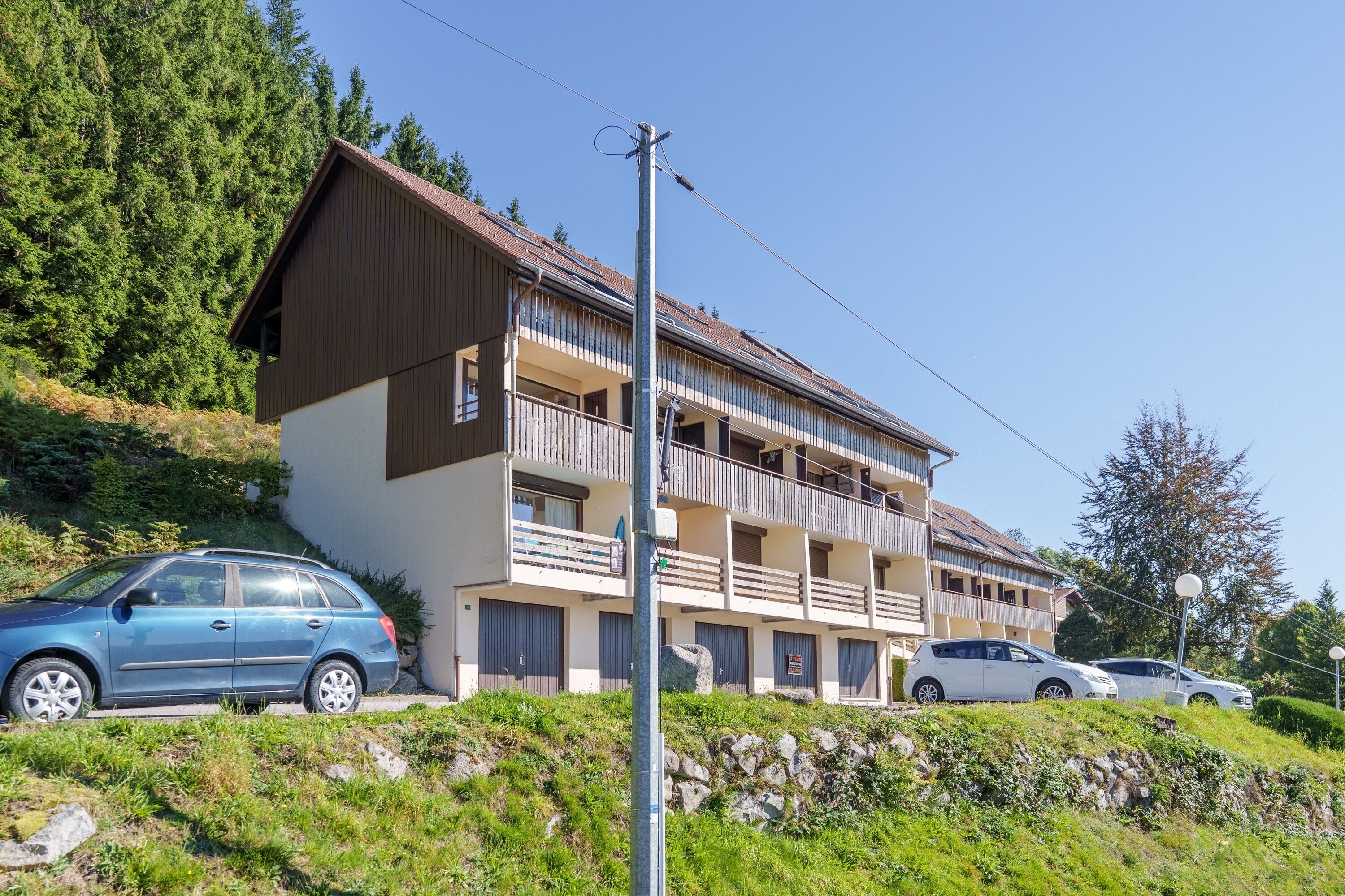 A multi-story building with balconies, parked cars, and a clear sky.