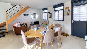 A modern living room with a wooden dining table, comfortable seating, a staircase, and a large window with blinds. - Gîte des Mouettes - Avec terrasse (Ghyvelde)