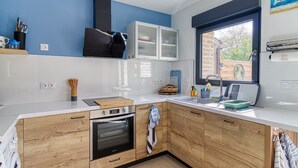 A modern kitchen with wooden cabinets, a built-in oven, a window with a view of the outside, and a countertop with various kitchen items. - Gîte des Mouettes - Avec terrasse (Ghyvelde)