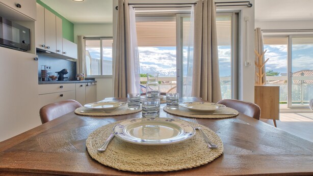 Une table à manger dressée avec des assiettes, des couverts et des verres, offrant une vue sur le ciel à travers de grandes fenêtres.