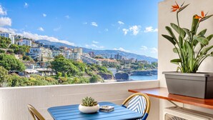 A balcony with a table and chairs, a potted plant, and a view of a coastal cityscape.