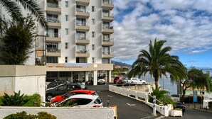 A multi-story residential building with balconies, a parking area with several cars, and a view of the sea.