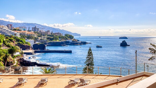 A coastal view from a balcony with lounge chairs, a rocky shoreline, and a distant cityscape.