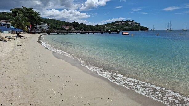 A beach with clear water, a pier, and houses in the background.