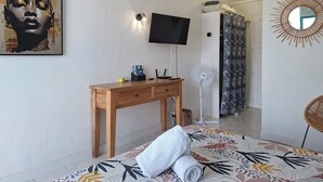 A hotel room with a bed, a wooden desk, a TV, a fan, and a wall-mounted artwork.
