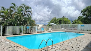 A swimming pool with stone tiles, a white fence, and palm trees in the background.