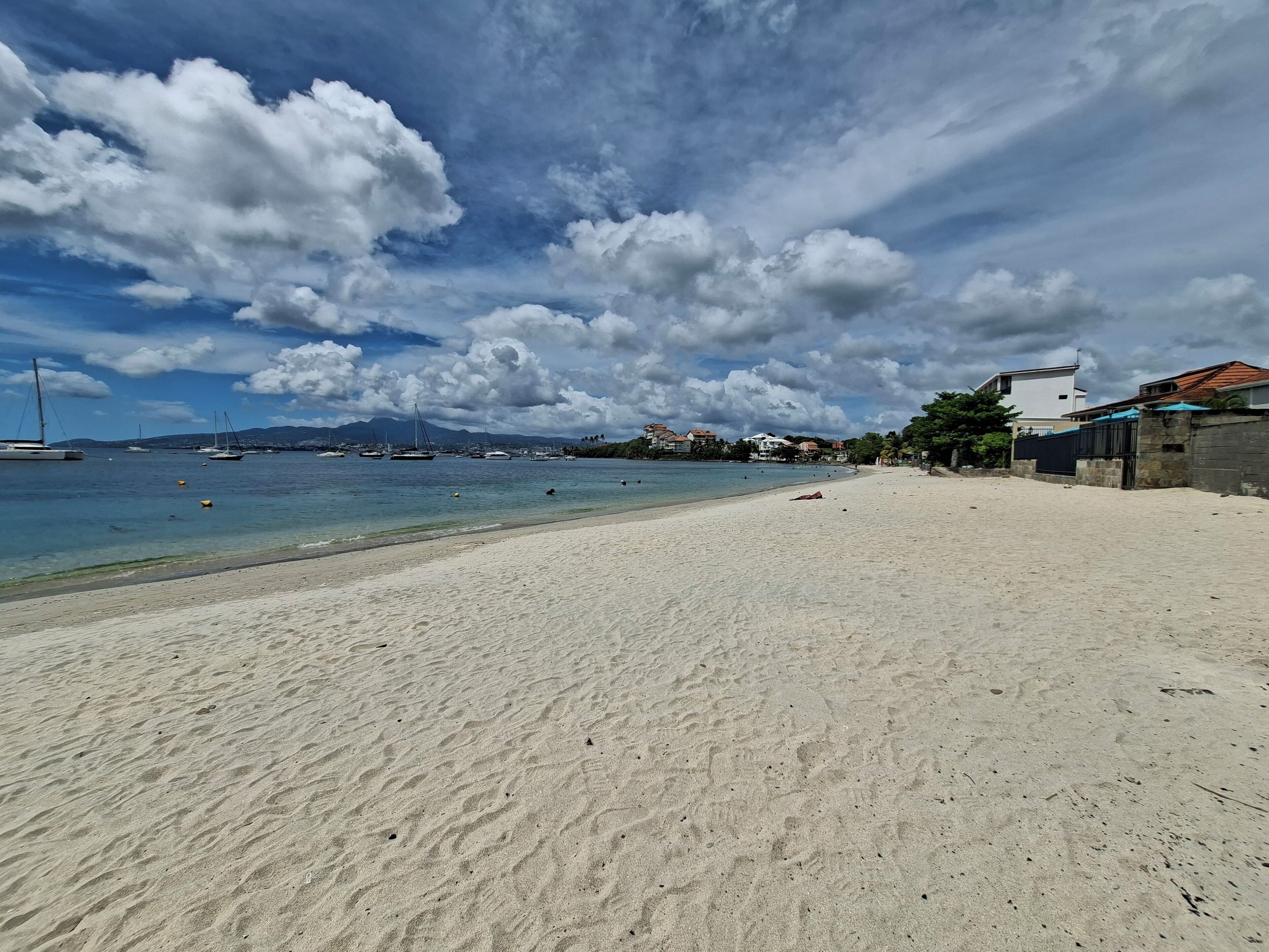 A beach with clear water, boats, and buildings in the background.
