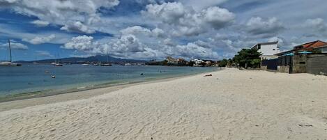 A beach with clear water, boats, and buildings in the background.