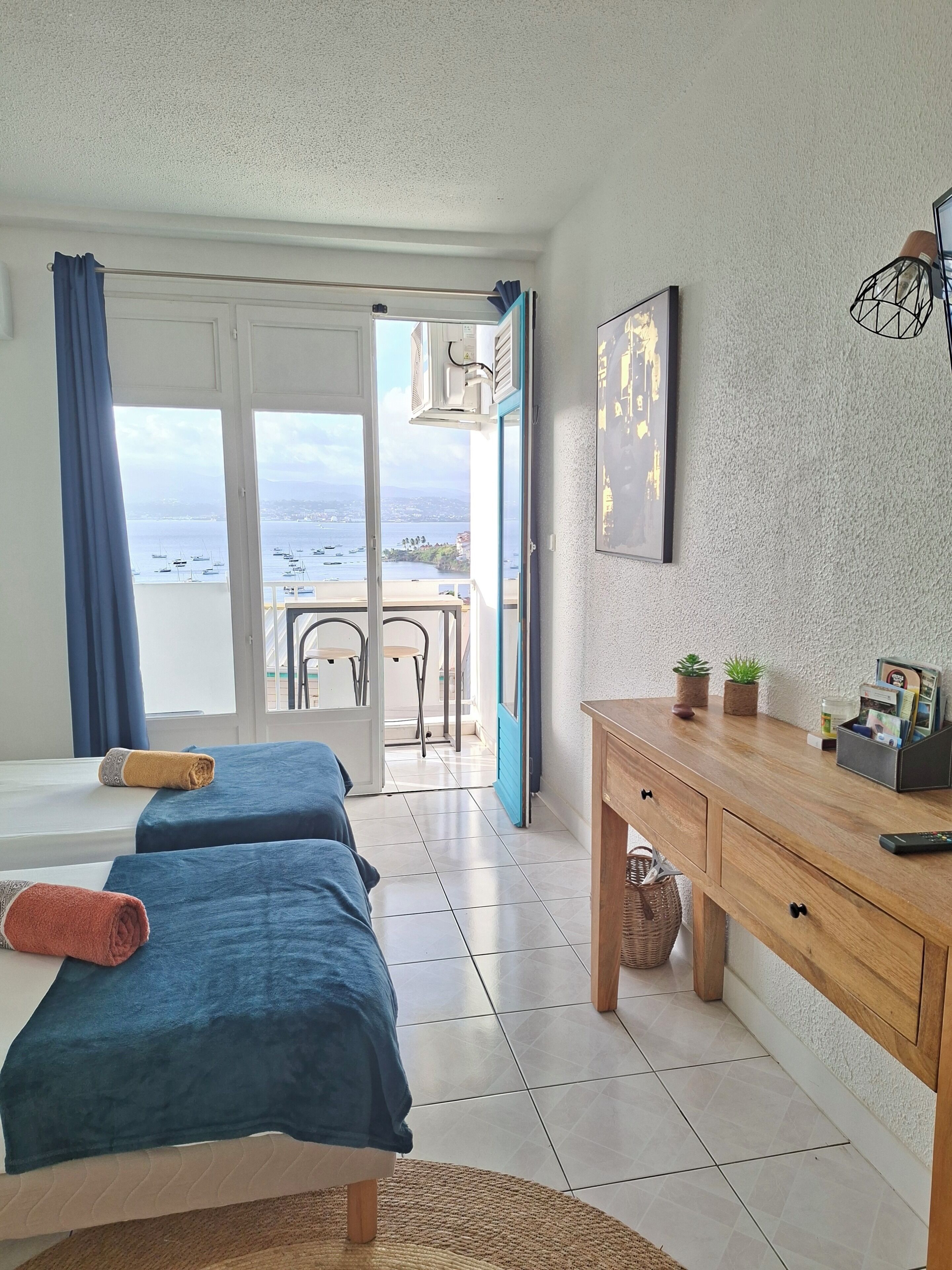 Une chambre d’hôtel avec balcon, deux lits avec des serviettes, un bureau en bois et une vue sur la mer.