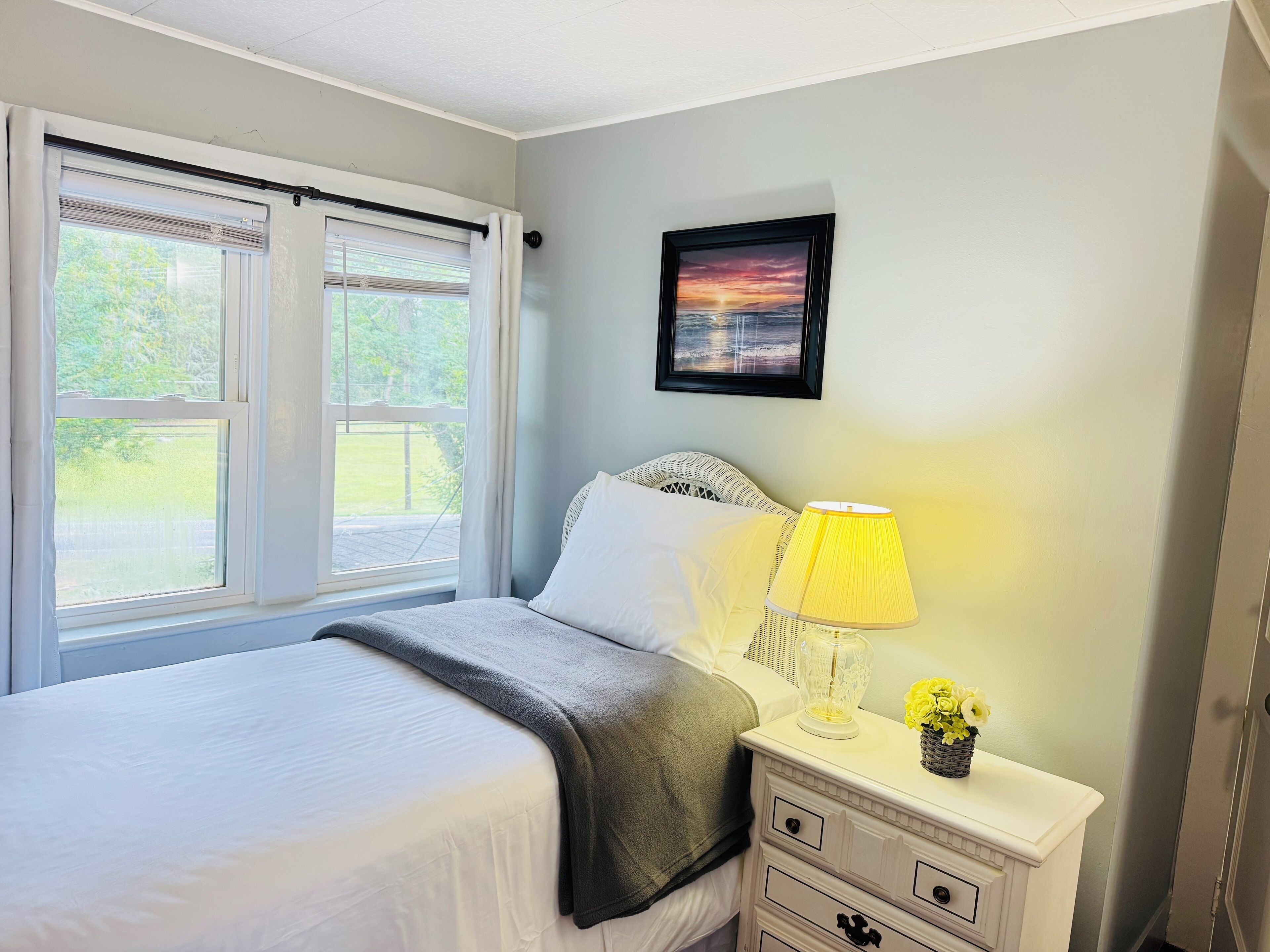 A bedroom with a bed, a bedside table with a lamp, a framed picture on the wall, and a window with curtains.