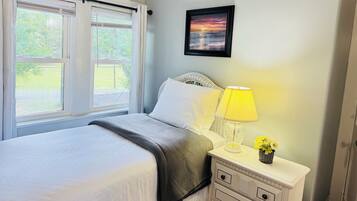 A bedroom with a bed, a bedside table with a lamp, a framed picture on the wall, and a window with curtains.