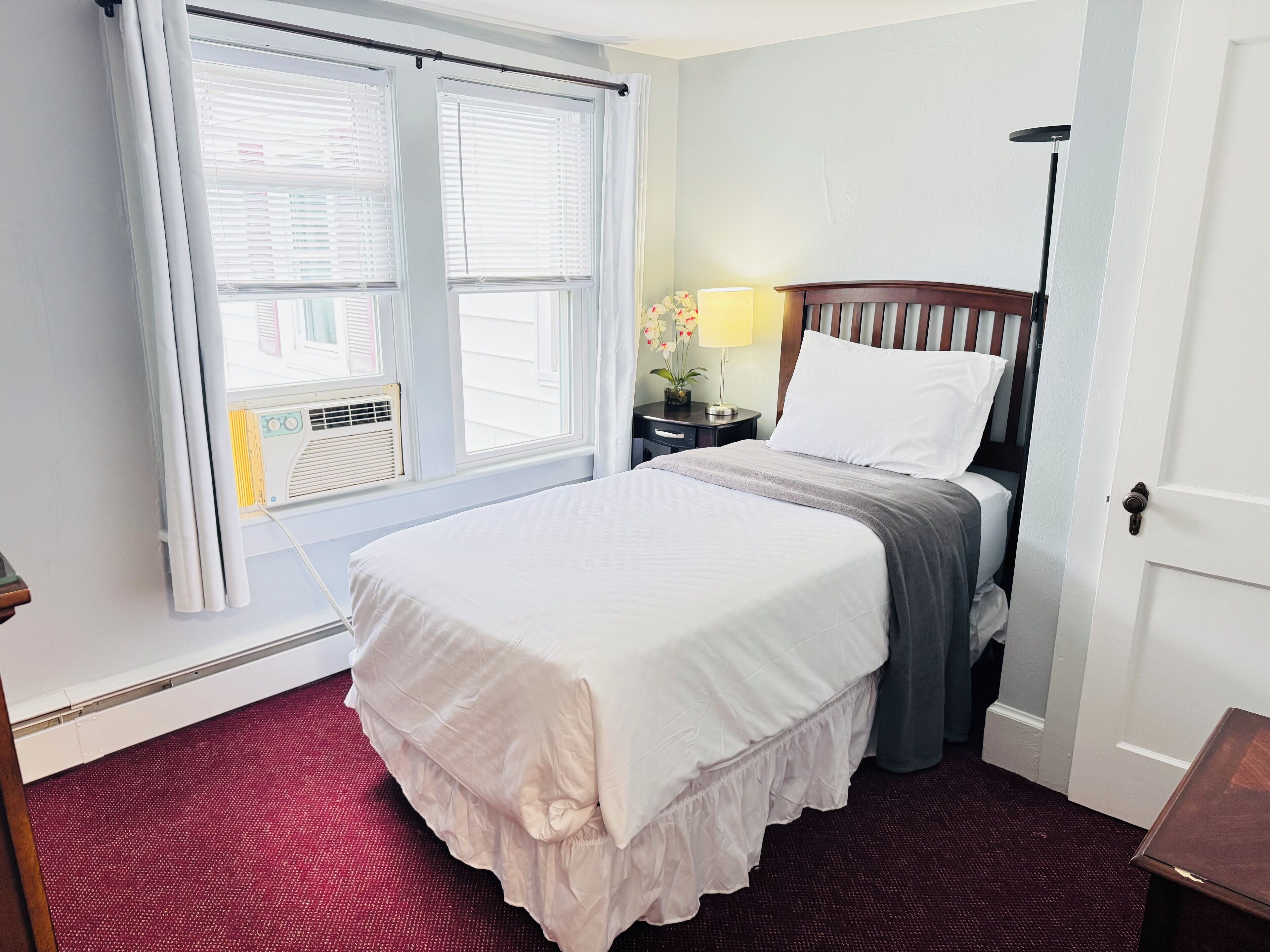 A neatly arranged hotel room with a bed, a nightstand, a lamp, and a window with blinds.