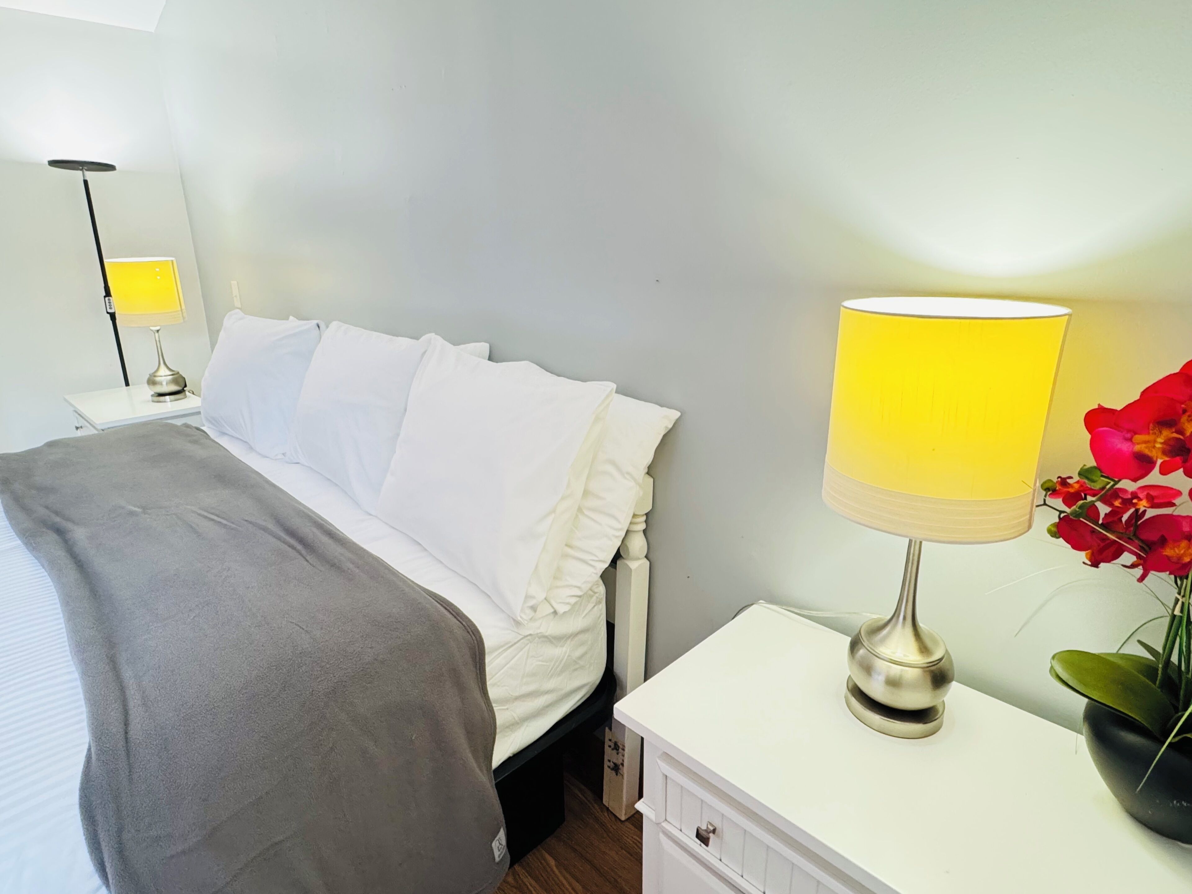 A neatly made bed with white linens and a grey blanket, a bedside lamp with a yellow shade, a small bedside table with a black lamp, and a potted plant with red flowers.