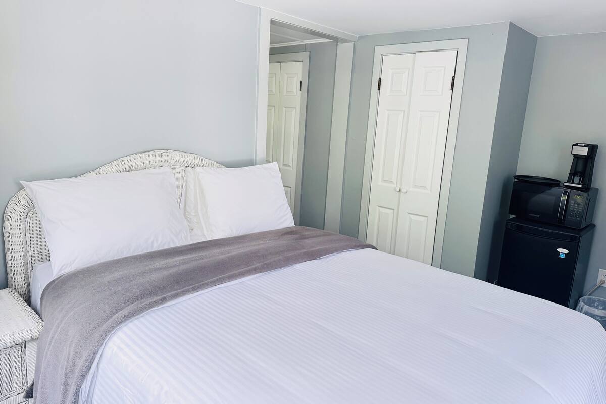 A neatly made bed with white linens and a gray throw blanket, a wicker chair, and a black coffee maker on a cabinet.