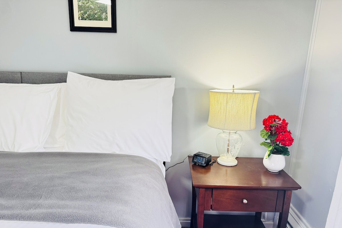 A neatly made bed with white linens, a bedside table with a lamp and a vase of red flowers, and a framed picture on the wall.