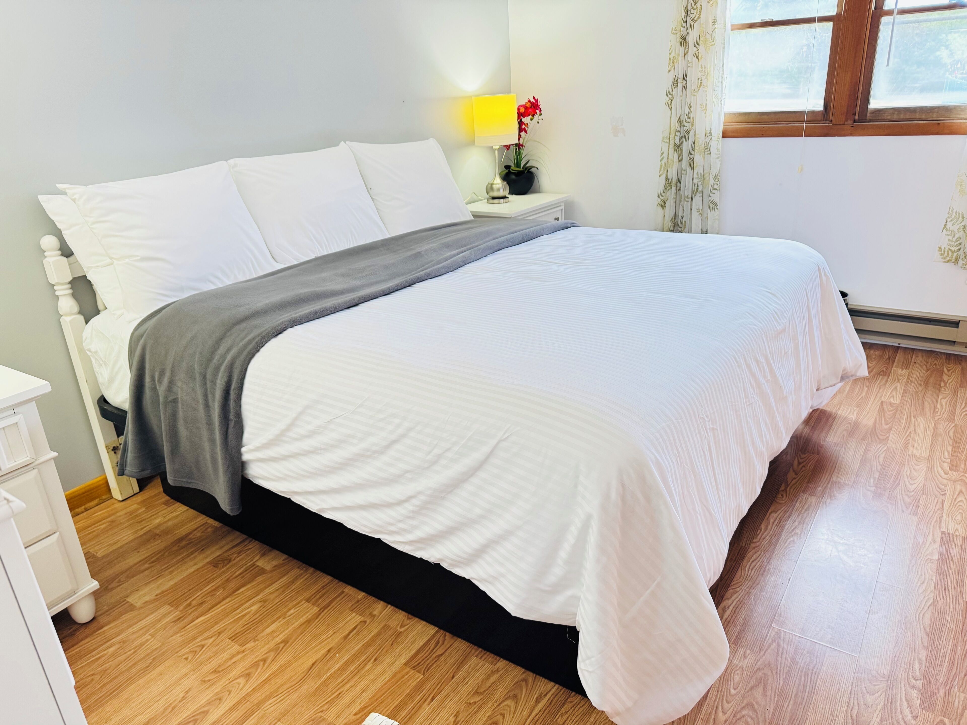 A neatly made bed with white and gray bedding, a bedside table with a lamp and flowers, and a window with curtains.