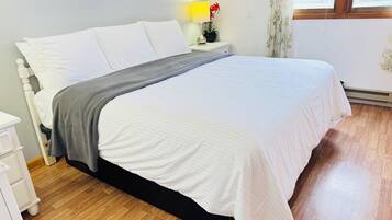 Una cama bien hecha con ropa de cama blanca y gris, una mesita de noche con una lámpara y flores, y una ventana con cortinas.