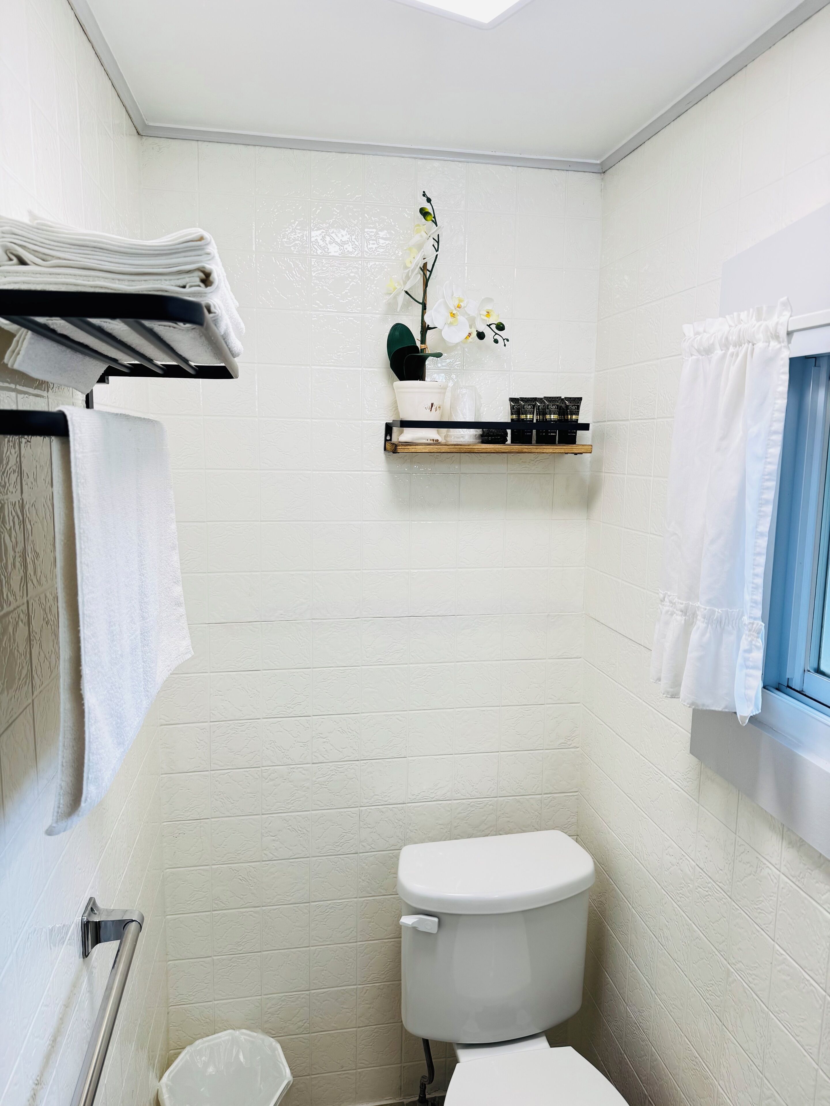 Un baño pequeño con paredes de azulejos blancos, un inodoro blanco, un estante con artículos de aseo y una ventana con cortina blanca.