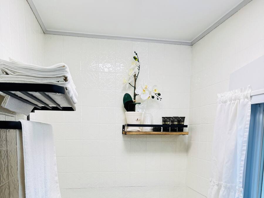 Un baño pequeño con paredes de azulejos blancos, un inodoro blanco, un estante con artículos de aseo y una ventana con cortina blanca.