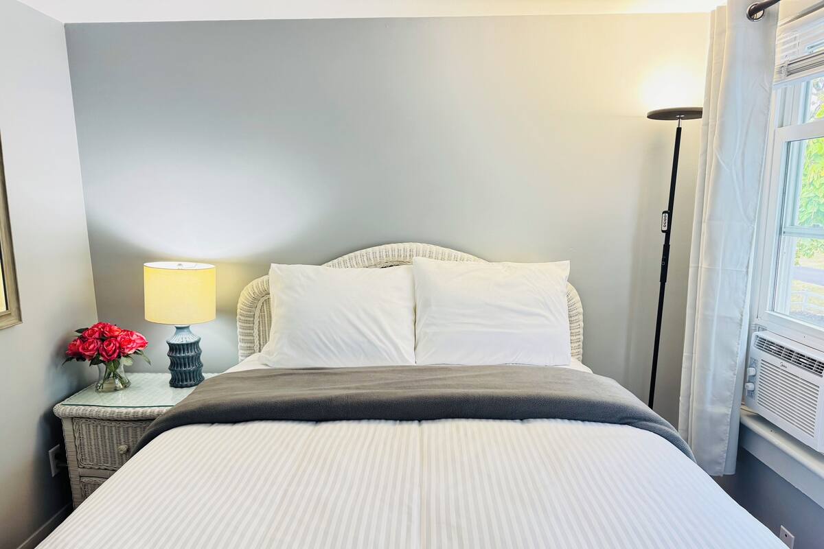 A neatly made bed with white bedding, a bedside lamp, and a vase of red flowers on a nightstand.