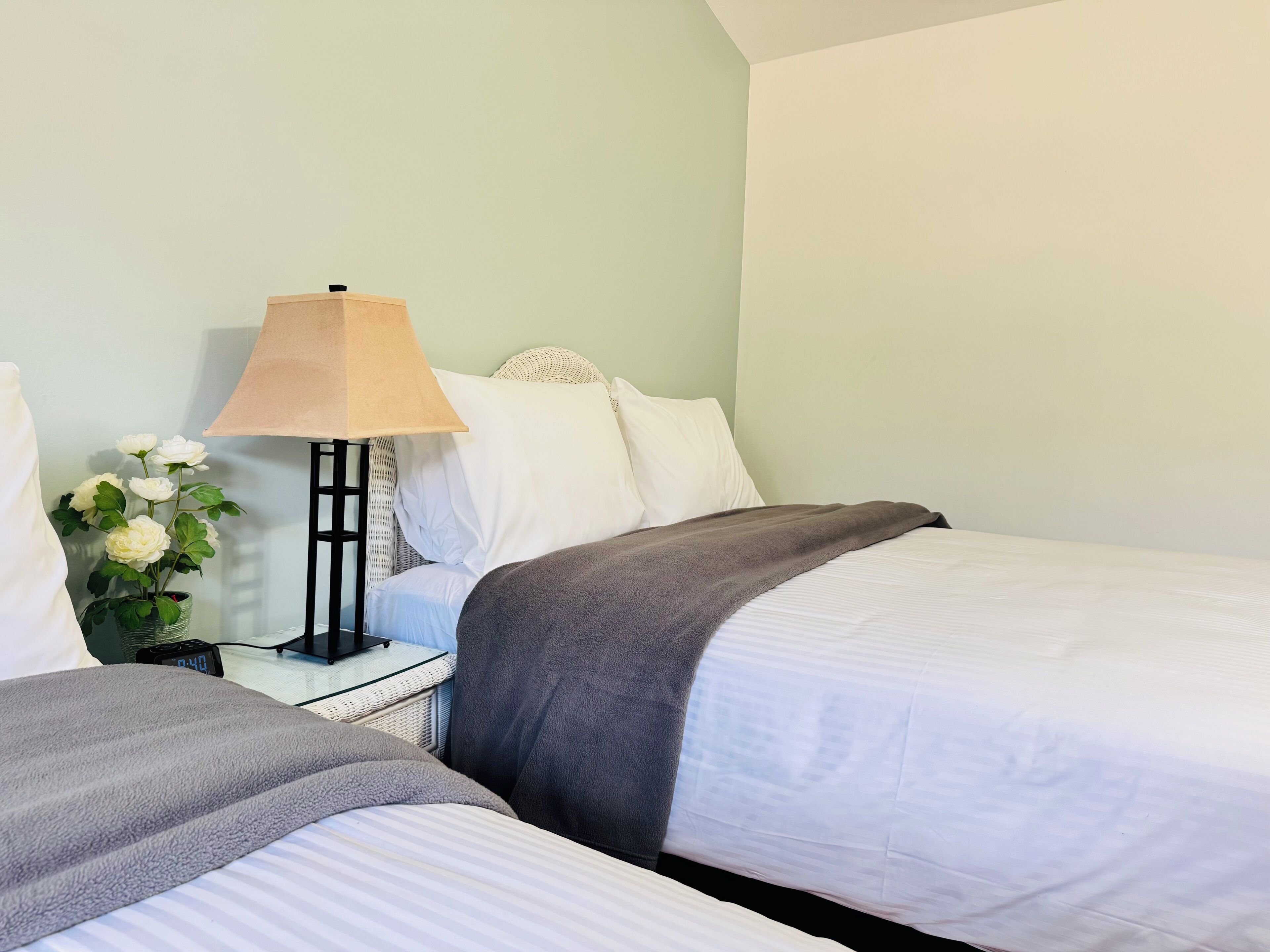 A neatly made twin bed with a gray blanket, a bedside table with a lamp, and a vase with white flowers.