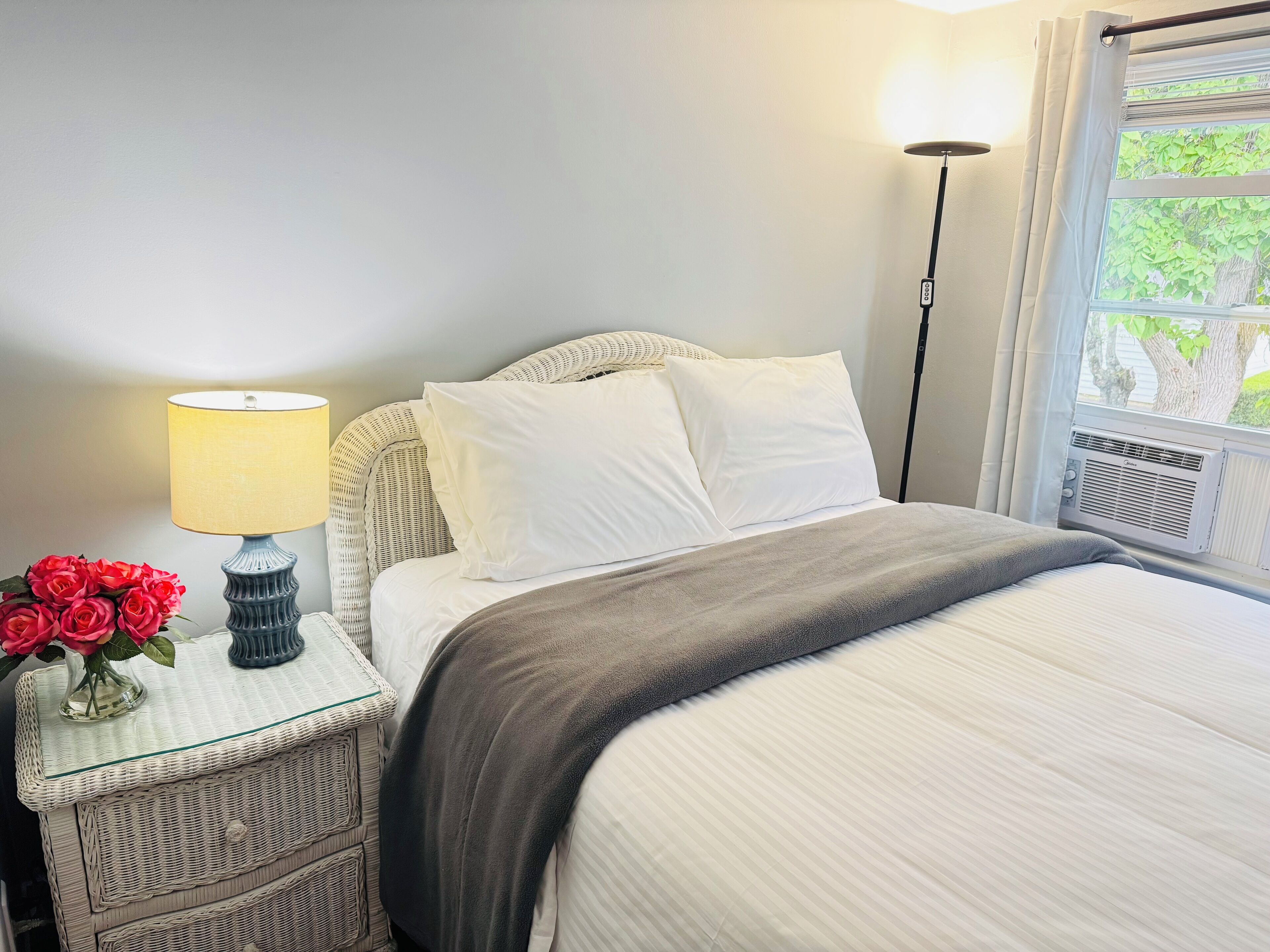 A neatly made bed with white linens, a grey throw blanket, and a bedside table with a lamp and a vase of red roses.