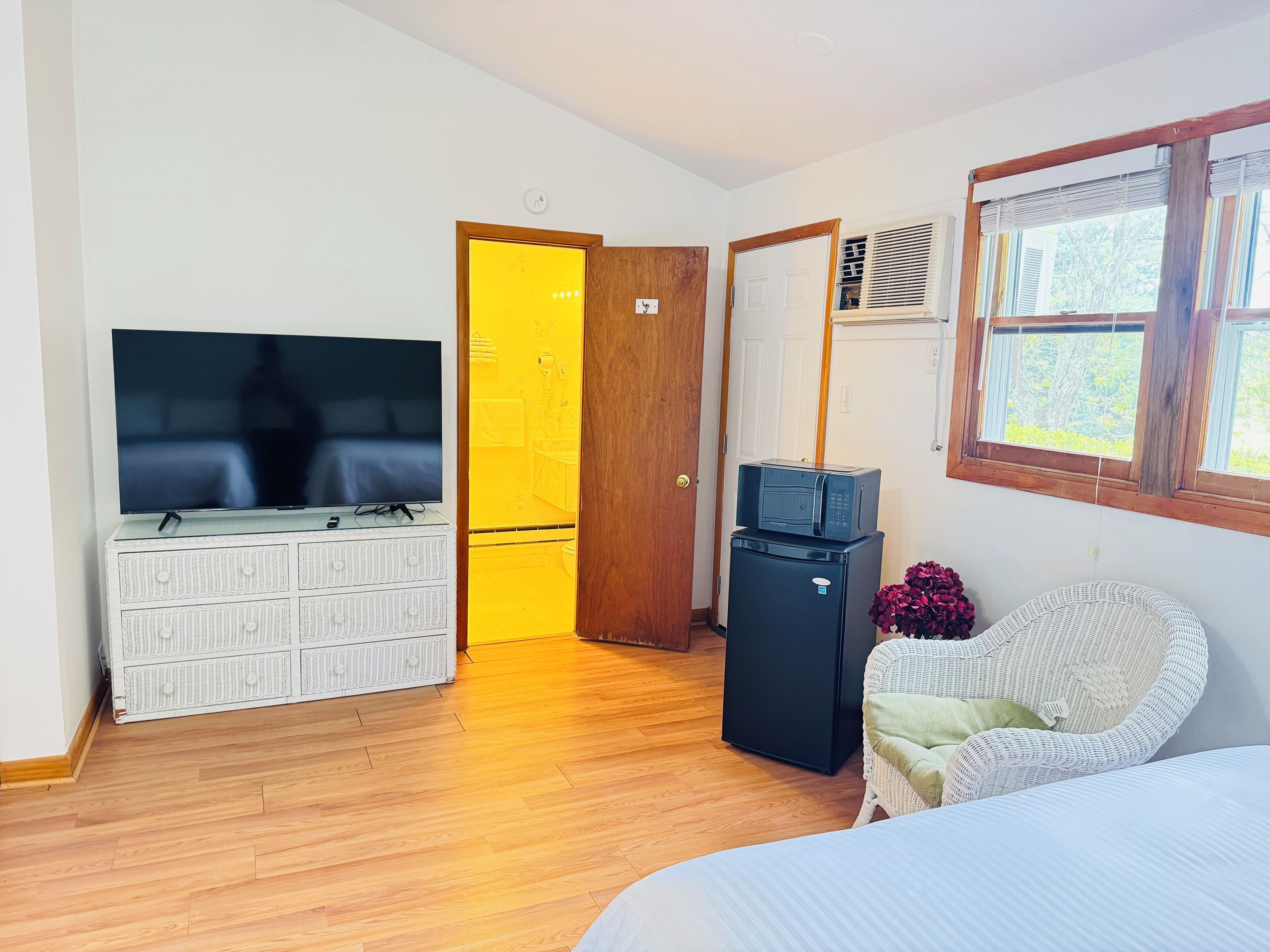 A room with a bed, a TV on a white dresser, a microwave on a black cabinet, a wicker chair, and a window with blinds.