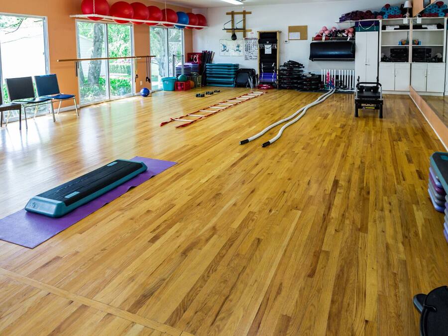 A spacious gym with wooden floors, exercise equipment, and a large window providing natural light.