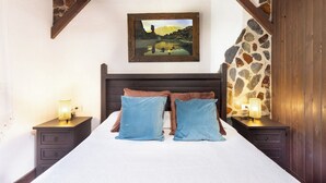 Une chambre à coucher avec une tête de lit en bois, deux tables de chevet avec des lampes, un lit avec des oreillers bleus et bruns, et un tableau représentant un paysage accroché au mur.
